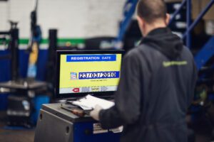 Vehicle tester in navy overalls holding a notepad inputs details on a computer inside a blue and white garage